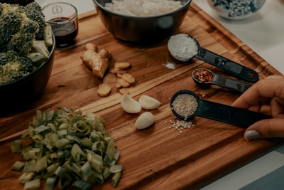A wooden cutting board displays various fresh ingredients including chopped green onions, garlic cloves, ginger, and broccoli in a bowl. Measuring spoons hold sesame seeds, crushed red pepper, and flour. A hand is holding the spoon with sesame seeds. A small bowl with soy sauce is also visible.