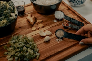 A wooden cutting board displays various fresh ingredients including chopped green onions, garlic cloves, ginger, and broccoli in a bowl. Measuring spoons hold sesame seeds, crushed red pepper, and flour. A hand is holding the spoon with sesame seeds. A small bowl with soy sauce is also visible.