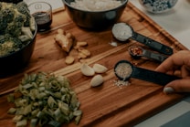 A wooden cutting board displays various fresh ingredients including chopped green onions, garlic cloves, ginger, and broccoli in a bowl. Measuring spoons hold sesame seeds, crushed red pepper, and flour. A hand is holding the spoon with sesame seeds. A small bowl with soy sauce is also visible.