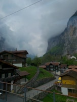 A picturesque mountain village with traditional wooden chalets nestled in a lush green valley. Majestic cliffs rise sharply in the background, partially shrouded by clouds. A narrow winding path leads through the village, surrounded by rustic buildings and dense greenery.