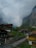 A picturesque mountain village with traditional wooden chalets nestled in a lush green valley. Majestic cliffs rise sharply in the background, partially shrouded by clouds. A narrow winding path leads through the village, surrounded by rustic buildings and dense greenery.