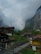 A picturesque mountain village with traditional wooden chalets nestled in a lush green valley. Majestic cliffs rise sharply in the background, partially shrouded by clouds. A narrow winding path leads through the village, surrounded by rustic buildings and dense greenery.