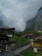 A picturesque mountain village with traditional wooden chalets nestled in a lush green valley. Majestic cliffs rise sharply in the background, partially shrouded by clouds. A narrow winding path leads through the village, surrounded by rustic buildings and dense greenery.