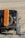 A rugged off-road vehicle is equipped with an orange traction board mounted on the spare tire at the back. The vehicle is positioned on a rocky, uneven terrain with a dry, barren landscape in the background.