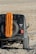 A rugged off-road vehicle is equipped with an orange traction board mounted on the spare tire at the back. The vehicle is positioned on a rocky, uneven terrain with a dry, barren landscape in the background.