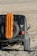 A rugged off-road vehicle is equipped with an orange traction board mounted on the spare tire at the back. The vehicle is positioned on a rocky, uneven terrain with a dry, barren landscape in the background.