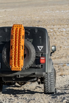 A rugged off-road vehicle is equipped with an orange traction board mounted on the spare tire at the back. The vehicle is positioned on a rocky, uneven terrain with a dry, barren landscape in the background.