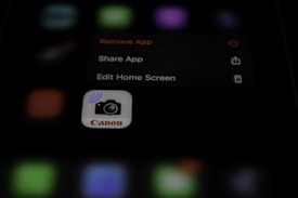 A blurred image with a focus on a smartphone screen displaying an app management menu. The options visible include 'Remove App', 'Share App', and 'Edit Home Screen'. An icon for a Canon camera app is also prominent.