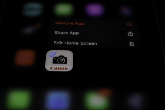 A blurred image with a focus on a smartphone screen displaying an app management menu. The options visible include 'Remove App', 'Share App', and 'Edit Home Screen'. An icon for a Canon camera app is also prominent.