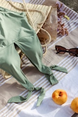 A green swimsuit is laid out on a striped towel next to a woven straw bag. Sunglasses are positioned nearby, and two peaches rest on the towel. Some dried flowers are partially visible, adding a natural element to the scene.