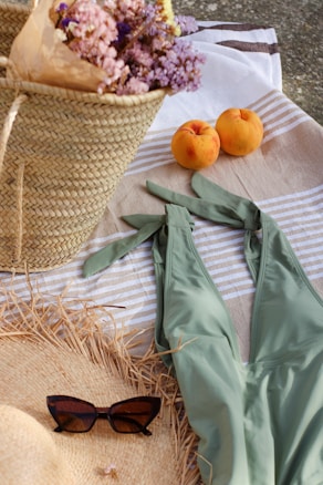 A woven straw basket filled with pastel-colored flowers sits on a striped beach towel next to two ripe peaches. A light green swimsuit is laid out alongside, with a pair of dark sunglasses resting on a straw sun hat.