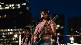 A woman is performing on stage with an electric guitar. She is wearing a light pink top with a floral pattern and blue jeans. The background reveals a cityscape at night with blurred lights from buildings, creating a lively urban atmosphere.