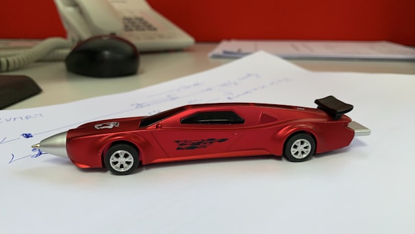 A sleek, red toy car with aerodynamic design sits on a desk. The car has a pointed front, black windows, and stylish decals on the side. It is placed on a white piece of paper with handwritten notes. In the background, there is an office telephone and other office items, slightly out of focus.