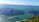 A large freight ship is sailing on a vast expanse of ocean under a clear blue sky. In the foreground, there are pieces of grass and plants, and in the distance, a landmass is visible on the horizon. Small waves can be seen on the water's surface.