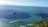 A large freight ship is sailing on a vast expanse of ocean under a clear blue sky. In the foreground, there are pieces of grass and plants, and in the distance, a landmass is visible on the horizon. Small waves can be seen on the water's surface.