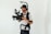 A person is operating a professional video camera mounted on a stabilizing rig. The individual is wearing a black hat and a white t-shirt, with tattoos visible on one arm. The background is plain and light-colored, creating a neutral setting.