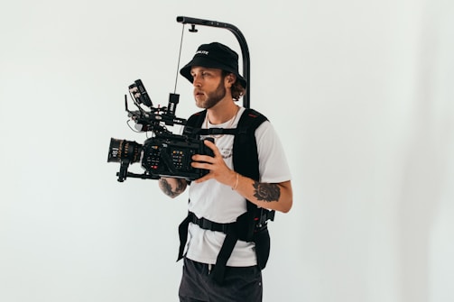 A person is operating a professional video camera mounted on a stabilizing rig. The individual is wearing a black hat and a white t-shirt, with tattoos visible on one arm. The background is plain and light-colored, creating a neutral setting.