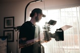 A person with a beard and a tattoo on the arm is holding a large, professional video camera. The individual is wearing a cap and carrying photography or videography equipment, with bright sunlight streaming through a window with blinds in the background.