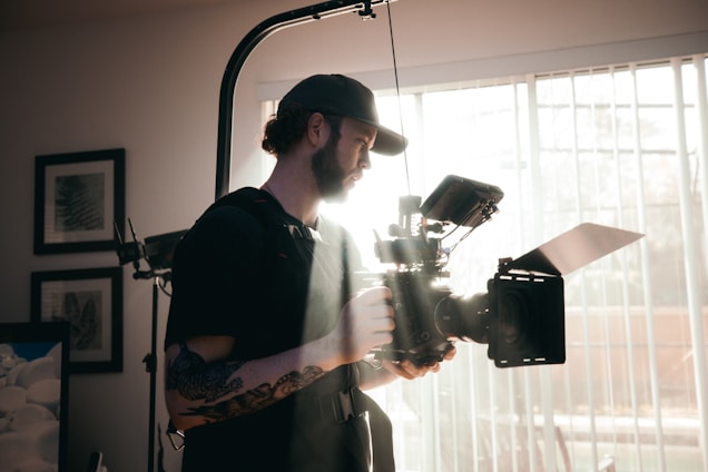 A person with a beard and a tattoo on the arm is holding a large, professional video camera. The individual is wearing a cap and carrying photography or videography equipment, with bright sunlight streaming through a window with blinds in the background.