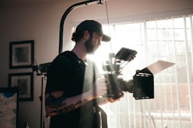 A person with a beard and a tattoo on the arm is holding a large, professional video camera. The individual is wearing a cap and carrying photography or videography equipment, with bright sunlight streaming through a window with blinds in the background.