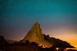 A tall, jagged rock formation is silhouetted against a starry night sky. The foreground is dimly illuminated, highlighting the rocky terrain and casting shadows. The sky features a gradient from deep blue at the top to a warm orange near the horizon.