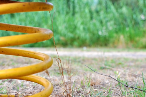 A large yellow coil spring is positioned diagonally in the foreground on a dry, grassy area. The background is out of focus, featuring a lush, green foliage scene, adding a contrast to the sharp focus of the spring.