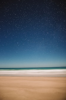 A serene beach scene under a clear, star-filled night sky. The gentle waves lightly touch the sandy shore, creating a peaceful and vast horizon.