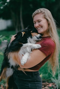 A woman with long blonde hair is smiling while holding a black-and-white dog. Both seem to be enjoying a peaceful moment outdoors, surrounded by greenery.