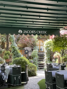 A charming outdoor dining area surrounded by lush greenery and plants. Tables are set with glassware and covered with dark green chair covers. A wooden staircase leads up to a rustic building with a small window at the top. An overhead awning with the logo 'Jacob's Creek' provides shade, while a large fan stands in the background.