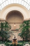 An indoor garden with a central stone fountain featuring a statue, surrounded by lush green foliage and flowering plants. Overhead, an arched glass ceiling allows natural light to illuminate the space. Elegantly designed architecture with beige walls and a hanging lantern adds to the tranquil and serene atmosphere.