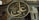 A large, circular ornate plaque with Arabic calligraphy is prominently displayed inside an architectural structure with intricate stone and decorative details. The background features arches and windows with metal grilles.