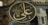 A large, circular ornate plaque with Arabic calligraphy is prominently displayed inside an architectural structure with intricate stone and decorative details. The background features arches and windows with metal grilles.