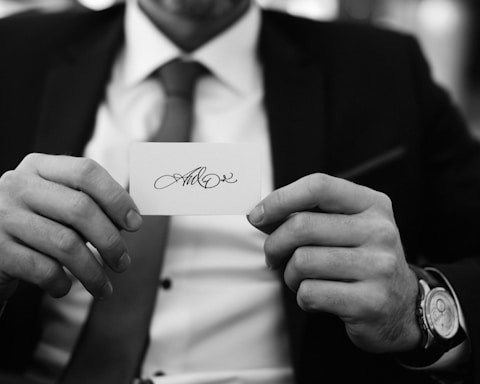 A person holding a business card with an elegant, cursive signature. The person is dressed in a suit, with a necktie and wearing a wristwatch. The background appears to be blurred.