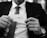 A person holding a business card with an elegant, cursive signature. The person is dressed in a suit, with a necktie and wearing a wristwatch. The background appears to be blurred.