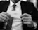 A person holding a business card with an elegant, cursive signature. The person is dressed in a suit, with a necktie and wearing a wristwatch. The background appears to be blurred.