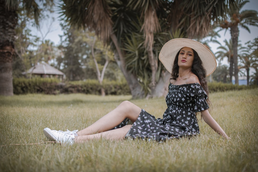 woman in black and white floral dress wearing brown sun hat sitting on green grass field