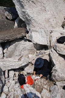 A person wearing a dark jacket and a blue hat is exploring a rocky area with large, white stones. They are using their hands to examine or move the rocks and nearby is a black backpack and some red fabric.