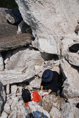 A person wearing a dark jacket and a blue hat is exploring a rocky area with large, white stones. They are using their hands to examine or move the rocks and nearby is a black backpack and some red fabric.