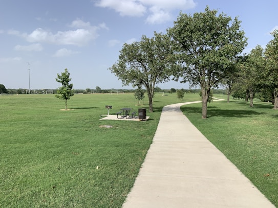 A spacious park features a winding concrete path flanked by well-maintained green grass and numerous trees. In the foreground, a picnic table with attached benches and a nearby grill sit on a small paved area. The sky is partly cloudy, creating a serene and inviting outdoor space.