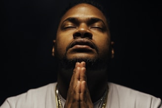 A man in a contemplative pose with hands in prayer position, eyes closed. The lighting is subdued, creating a serene and introspective ambiance. He is wearing a light-colored shirt and some necklaces that catch the dim light.