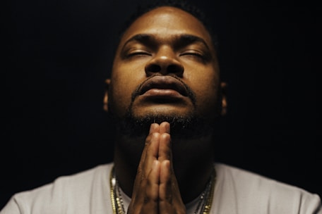 A man in a contemplative pose with hands in prayer position, eyes closed. The lighting is subdued, creating a serene and introspective ambiance. He is wearing a light-colored shirt and some necklaces that catch the dim light.