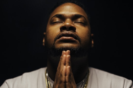 A man in a contemplative pose with hands in prayer position, eyes closed. The lighting is subdued, creating a serene and introspective ambiance. He is wearing a light-colored shirt and some necklaces that catch the dim light.