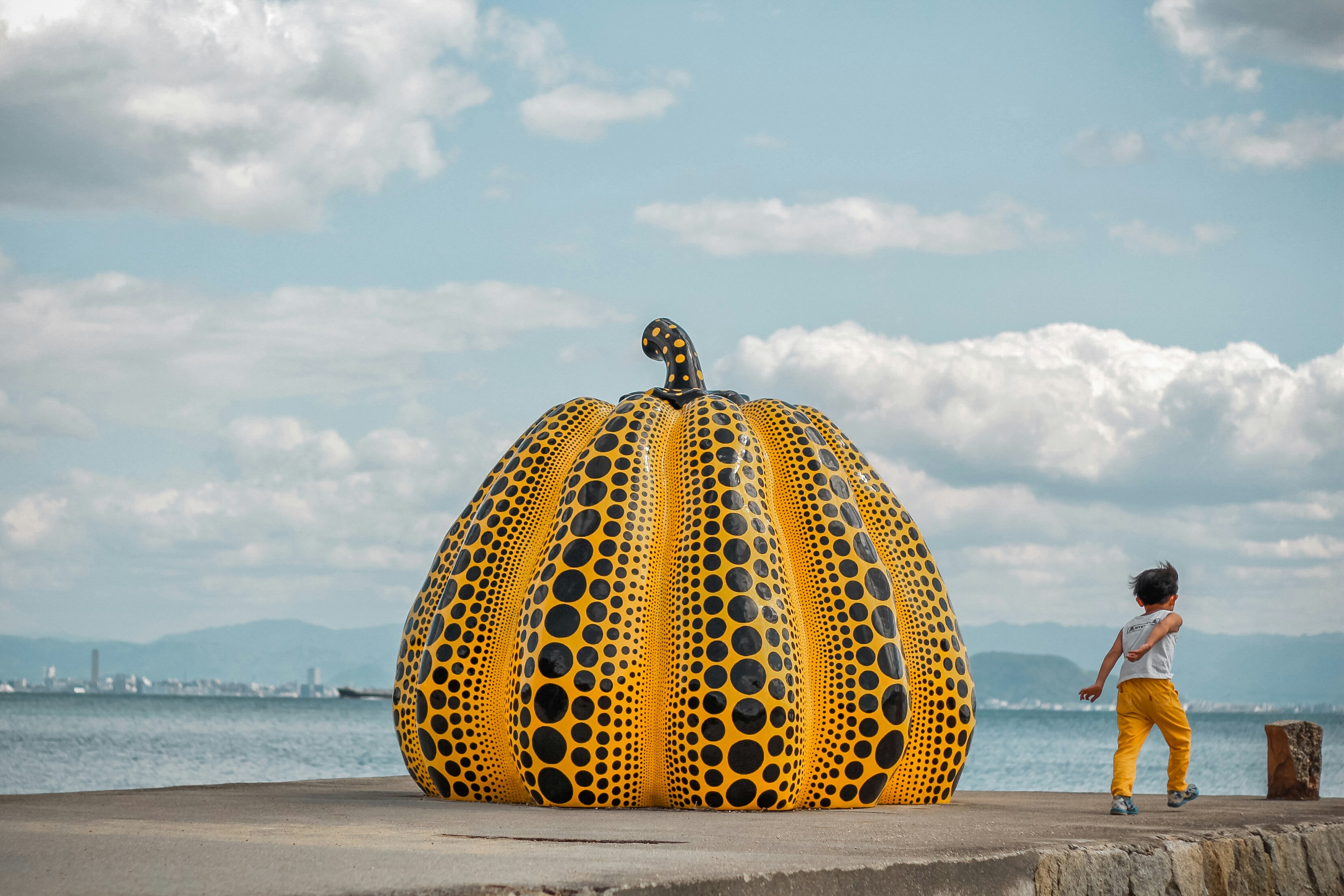 Image shows an artwork by Yayoi Kusama in the shape of a giant pumpkin speckled with black spots. A figure in white and orange stands with their back to the artwork. They are outside. 