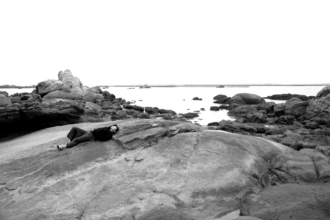 grayscale photo of man lying on rock near body of water