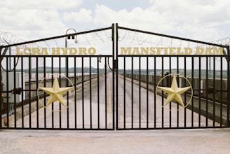 A metal gate with the words 'LCRA Hydro' and 'Mansfield Dam' in yellow lettering spans the width of the entrance. The gate features large yellow stars on each side, and the background reveals a paved pathway leading further back, likely for maintenance or restricted access. Barbed wire tops the fence, indicating a secure area. The sky is partly cloudy, and there are hints of a water body and distant landscape.