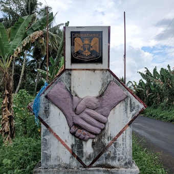 A concrete monument features an emblem with an eagle and the word 'minahasa' at the top. Below, there's a large sculpture of two hands engaging in a handshake. The monument is surrounded by lush greenery and banana trees, with a road running by its side. The sky is partly cloudy, suggesting a pleasant and calm setting.