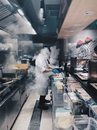 In a bustling kitchen, a chef wearing a white uniform and blue gloves is preparing food amidst steam and smoke. Stainless steel counters lined with various containers and utensils surround the workspace. Another person in the background is partially visible, adding to the busy ambiance of the kitchen. The wall features artistic fish drawings, enhancing the aesthetic.