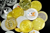 A pile of various cryptocurrency coins is scattered on a surface. The coins display different designs and logos, with a mix of gold and silver colors. Notable graphics include a Shiba Inu image and geometric shapes, representing popular cryptocurrencies.