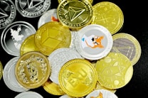 A pile of various cryptocurrency coins is scattered on a surface. The coins display different designs and logos, with a mix of gold and silver colors. Notable graphics include a Shiba Inu image and geometric shapes, representing popular cryptocurrencies.