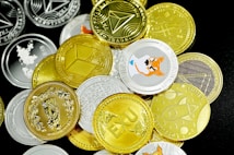 A pile of various cryptocurrency coins is scattered on a surface. The coins display different designs and logos, with a mix of gold and silver colors. Notable graphics include a Shiba Inu image and geometric shapes, representing popular cryptocurrencies.